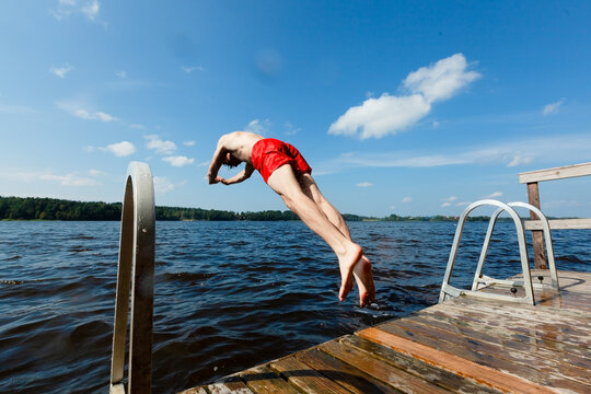 Guy dives off dock into calm lake in free fall on warm summer day. Young man in red swim trunks plunges with athletic freedom outdoors. Diver descends through air creating splash entry in the water