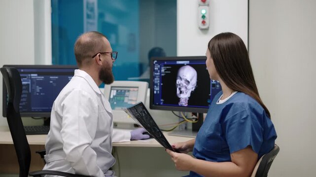 Radiologist and nurse review brain scan images on medical monitor, discussing MRI results and diagnostic findings in hospital office.