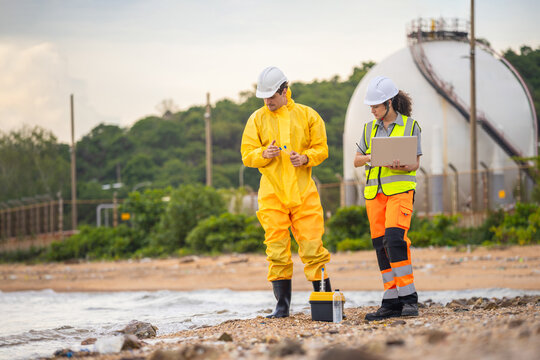 Environmental scientists in protective gear testing water quality at industrial site, Professional team performing hazardous material analysis and environmental assessment near oil refinery coastline
