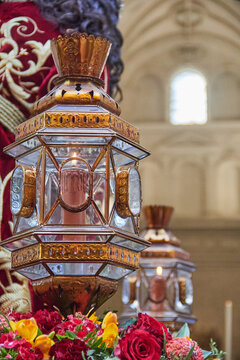 Ornate glass lantern with lit candle placed on a processional platform, surrounded by flowers and rich textiles, representing Catholic ceremonial lighting and devotional tradition