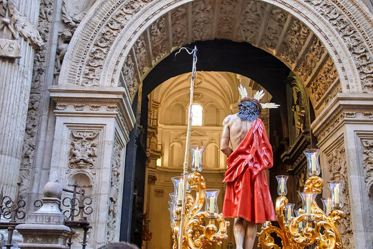Religious paso with sculpture of Jesus Christ in red robe entering cathedral doorway, framed by ornate architecture and candles, expressing Catholic devotion and ceremonial tradition