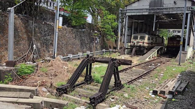 A railway yard featuring an Indian electric locomotive parked inside a maintenance shed. In the foreground, there are concrete sleepers and a black metal buffer stop at a track end.