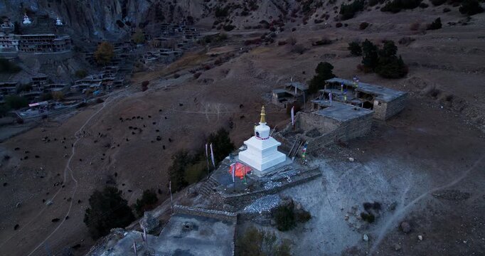 An aerial drone view captures a Buddhist stupa with golden spire standing among a remote Himalayan village nestled at the foot of rocky cliffs in Nepal.