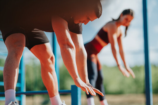 A man and woman stretch forward as they warm up for an outdoor workout. The pair perform standing hamstring stretches together at a sunny fitness park.