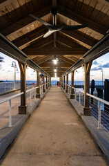 Obraz premium Wooden covered pier at dawn or sunset. Pier promenade deck leading to sea view. Seafront promenade with wood roof. Pier path on promenade avenue with nobody. Sea sunset horizon