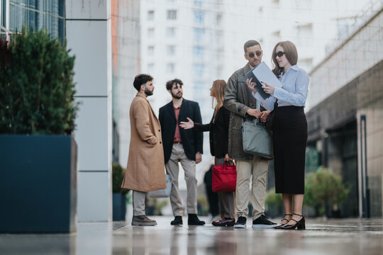 A group of business colleagues stand together on a city sidewalk, discussing documents. The professionals converse and review notes during an informal outdoor meeting in an urban setting.