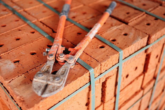 Heavy-duty bolt cutters with orange handles lying on a pallet of brand new red clay bricks secured with plastic strapping at a construction site.