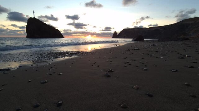 Sunset ocean beach, waves crashing sandy shore, dramatic cliffs, rocky island cross, peaceful tranquil evening seascape