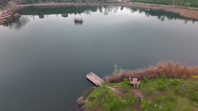 4K drone shot flying straight 20m above a calm lake from a dock toward the opposite shore. Cars pass on the road ahead under overcast skies with a cinematic, stable atmosphere.