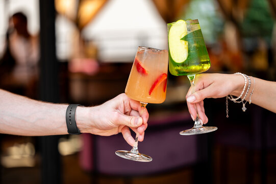 A close-up shot of two people clinking glasses with colorful summer cocktails garnished with fresh fruit at an outdoor restaurant.