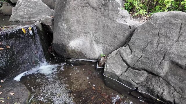 Slow motion of clear water flowing down from large rocks, a babbling brook and a small waterfall in a park