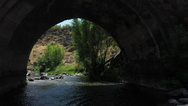 Camera moving forward under a beautiful medieval stone bridge arch over a fast, shallow river with small waterfalls, surrounded by lush green trees on a sunny day.

