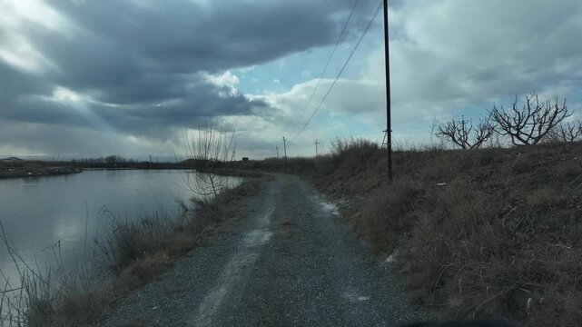 Camera moving forward on a gravel road next to a frozen lake on a cloudy winter day, with trees and electric poles on the side and mountains in the distance.

