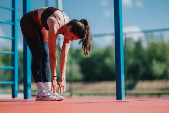 A young woman bends forward to stretch her hamstrings while standing on an outdoor sports court. She performs a pre-workout stretch in athletic clothing beside blue exercise bars.