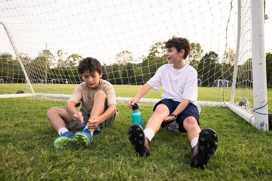 Two young boys sitting on the grassy field in front of soccer goal one fixing his shoe