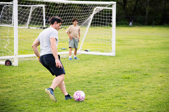 Adult male kicks a soccer ball on a grassy park field with young boy guarding the goal