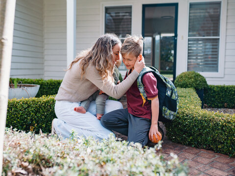 Australian mum kissing young son wearing backpack while kneeling on brick pathway in the front yard