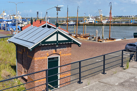 Den Oever, Netherlands - 20 May 2026: View of the historic former waterlevel gauge house at Vissershaven with fishing boats docked in the harbor under a clear blue sky.