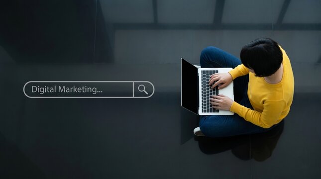 Overhead view of a woman in a yellow sweater working on a laptop with digital marketing text in a search bar on a black background banner