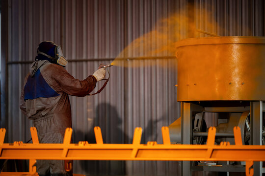 Industrial painter spray painting yellow coating on machinery metal parts, waring protective suit respirator mask spray painting yellow coating heavy machinery metal equipment factory workshop.