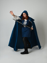 Naklejka premium full length portrait of female model wearing medieval fantasy warrior historical costume with blue hooded cloak. silhouette figure, holding sword weapon on studio background.