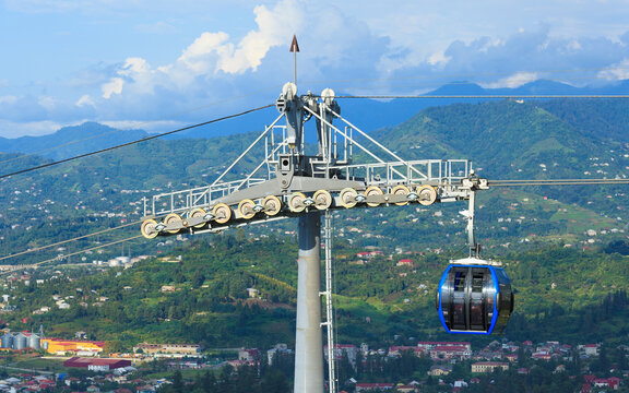 Modern cable car gondola suspended over mountainous landscape with green hills and residential areas below. Blue cabin hangs from steel cable system with mechanical pulleys visible on tower