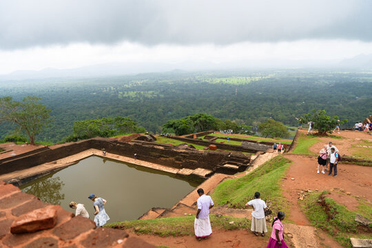 Sri Lanka - 21 February 2026: View of the ancient Sigiriya rock fortress ruins featuring a large water tank and tourists overlooking the vast green jungle landscape.