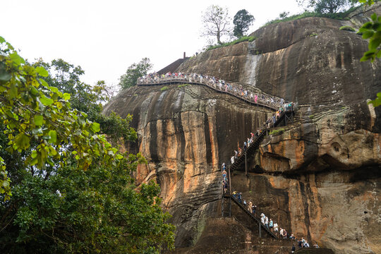 Sri Lanka - 21 February 2026: View of tourists climbing the steep metal staircase on the side of the ancient Sigiriya rock fortress with weathered rock face and lush tropical greenery.