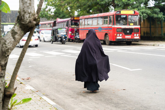 Sri Lanka - 15 February 2026: View of a woman in a black niqab walking on a sidewalk near a crosswalk with red buses and a tuk-tuk in the background.