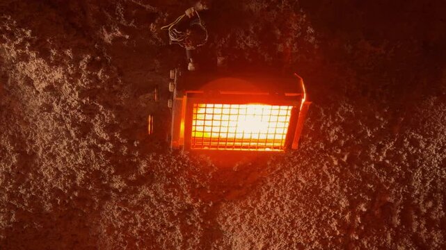 Industrial electric lamp fixture glowing orange behind metal grille mounted on rough rocky tunnel wall with water drip falling down, underground lighting scene