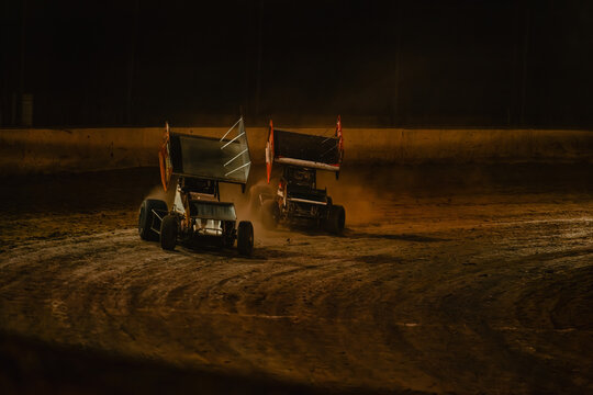 Sprint cars race on dirt track at night, kicking up dust as they round a bend