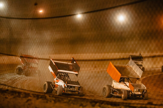 Sprint cars race on a dirt track at night kicking up dust under lights