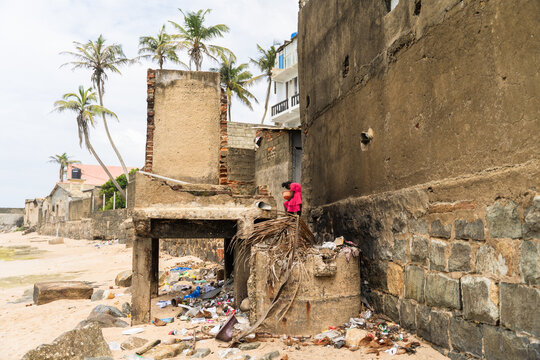 Sri Lanka - 05 February 2026: View of a woman in a pink dress standing by a weathered stone wall and debris on a sandy beach with palm trees.