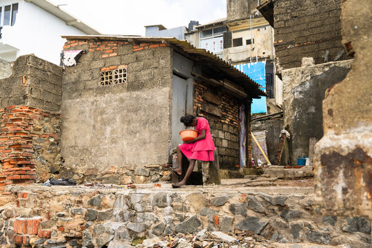 Sri Lanka - 05 February 2026: View of a young girl in a pink dress sitting outside a weathered brick building holding an orange bowl in a poor urban area.