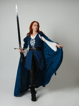 full length portrait of female model wearing medieval fantasy warrior historical costume with blue cloak. silhouette figure, holding sword fighting weapon on studio background.