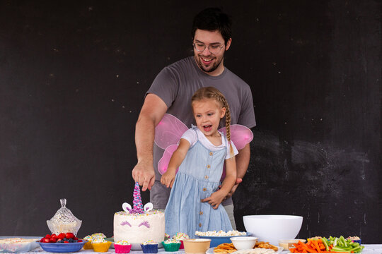 Childs unicorn birthday cake on table amongst party food with dad and little girl