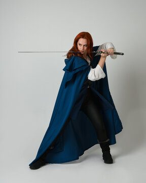 full length portrait of female model wearing medieval fantasy warrior historical costume with blue cloak. silhouette figure, holding sword fighting weapon on studio background.