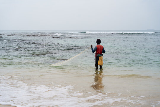 Sri Lanka - 03 February 2026: View of a fisherman in a red shirt standing in the shallow turquoise water of the Indian Ocean holding a casting net on a cloudy day.