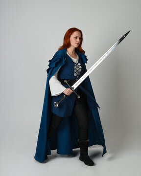 full length portrait of female model wearing medieval fantasy warrior historical costume with blue cloak. silhouette figure, holding sword fighting weapon on studio background.