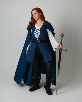 full length portrait of female model wearing medieval fantasy warrior historical costume with blue cloak. silhouette figure, holding sword fighting weapon on studio background.