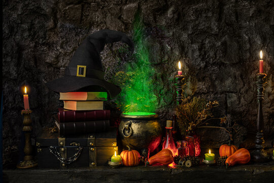 Halloween background. Dark witch cauldron with a magic witch's potion, books, chest, conical hat. Witchcraft paraphernalia and accessories among candles on a table in a room or cave with stone walls.