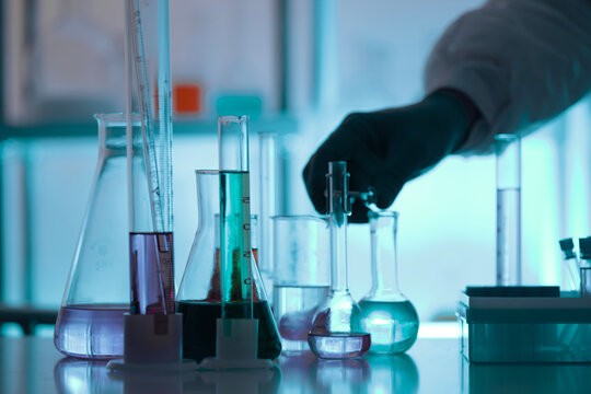 Chemistry laboratory scene with colorful chemical solutions in glass flasks and cylinders, scientist in gloves writing results, analytical experiment workspace.