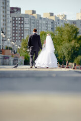Obraz premium A newly married couple in formal wedding attire strolls away along a city park path, with urban buildings and green trees in the background, symbolizing love, new beginnings, and romance.