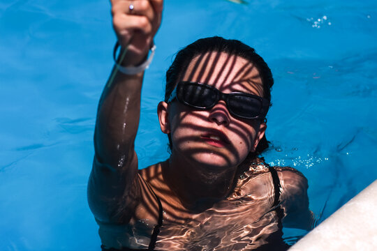 Woman in swimming pool raising arm, wearing sunglasses, dramatic shadow patterns on face, water reflections and blue pool, summer leisure and relaxation concept, stylish outdoor lifestyle
