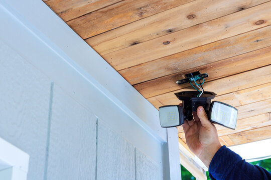 Handy man holds outdoor security lights while preparing to install them on wooden ceiling at their home.