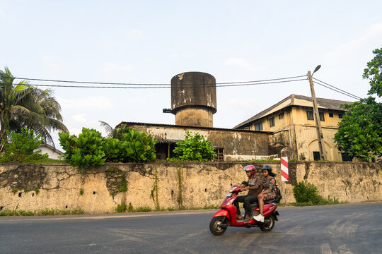 Sri Lanka - 15 January 2026: View of a man and a woman riding a red scooter past a weathered concrete water tower and historic stone walls.
