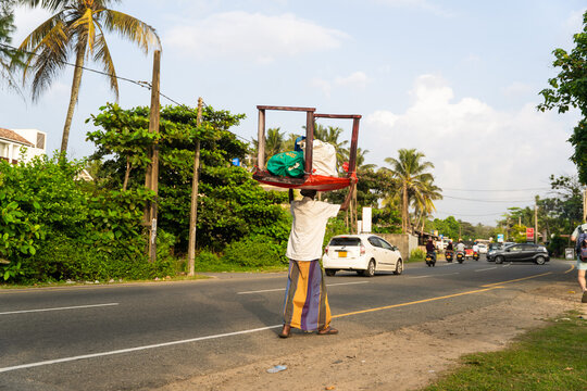 Sri Lanka - 15 January 2026: View of a man carrying a wooden table on his head while walking along a roadside with palm trees and traffic.