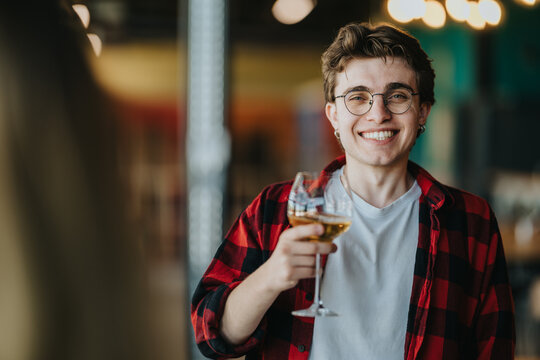 A smiling young man raises a glass of white wine while looking at the camera. He enjoys a casual indoor toast with friends at a relaxed social gathering.