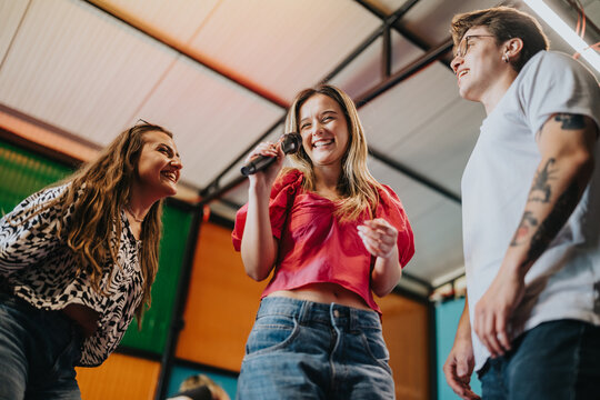 Three young friends sing into a microphone and laugh together while enjoying each other's company. The group shares a joyful moment performing and socializing in a relaxed indoor setting.