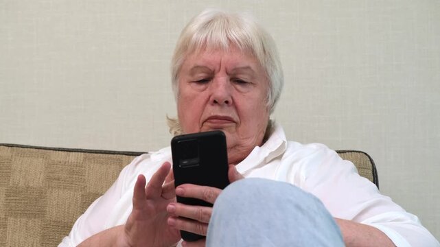 Focused senior woman using mobile phone while resting on couch close up. Concept of senior people adopting modern technology